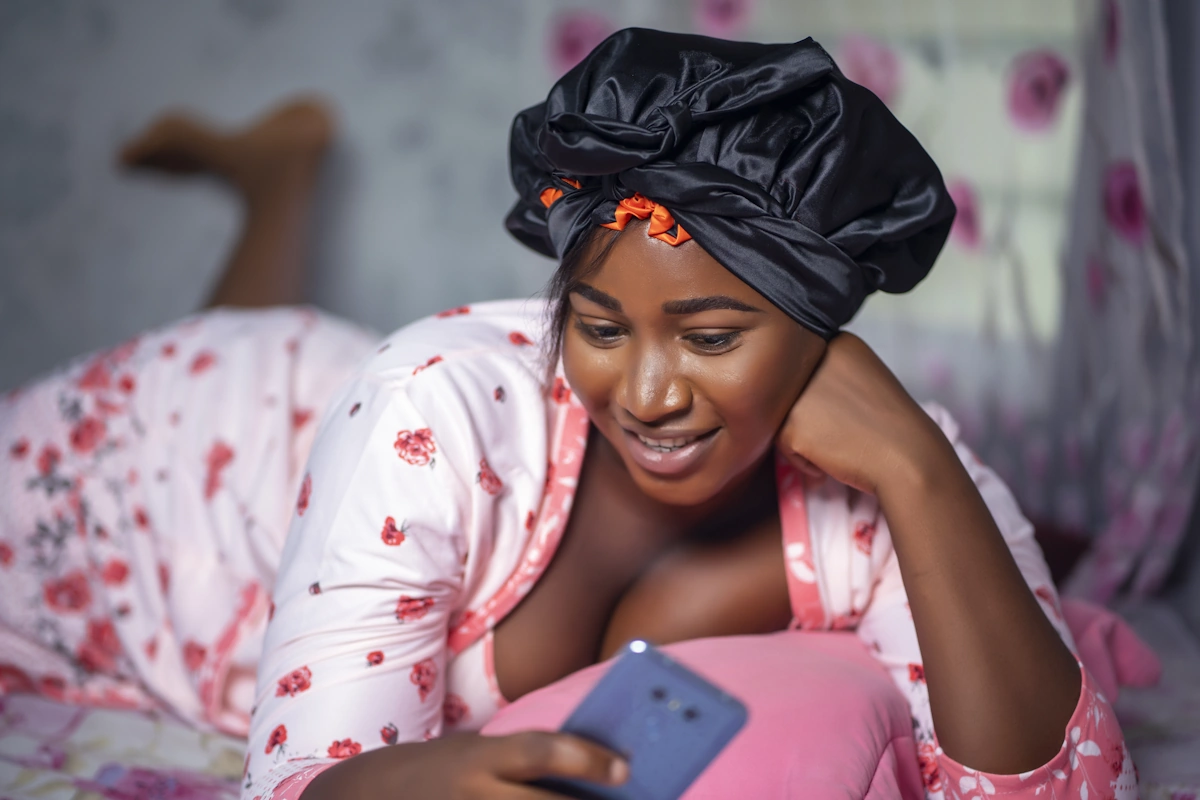 Une jeune femme avec un bonnet en satin qui joue sur son Smartphone sur le lit