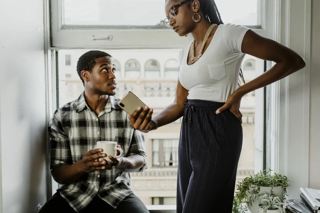 Une femme qui tend un téléphone à son mari qui la regarde assis la fenêtre une tasse à la main