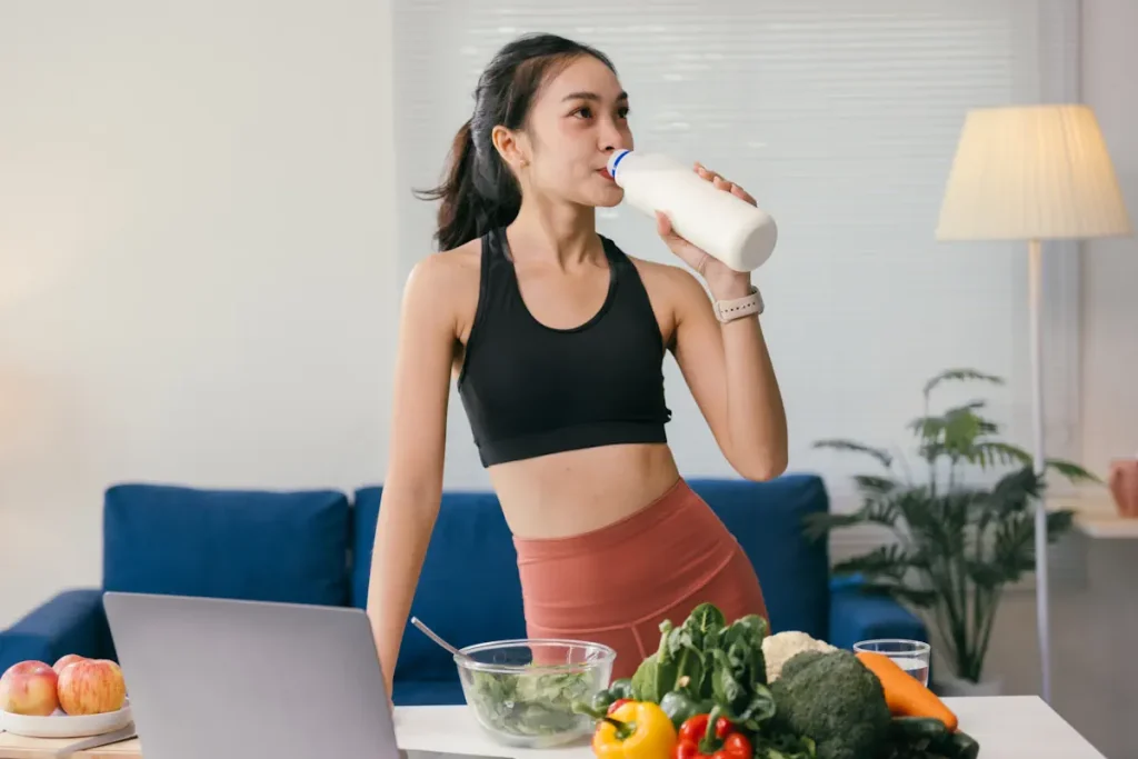 Une jeune femme asiatique qui boit dans une bouteille plastique blanche à côté d'une table où est posé différents aliments