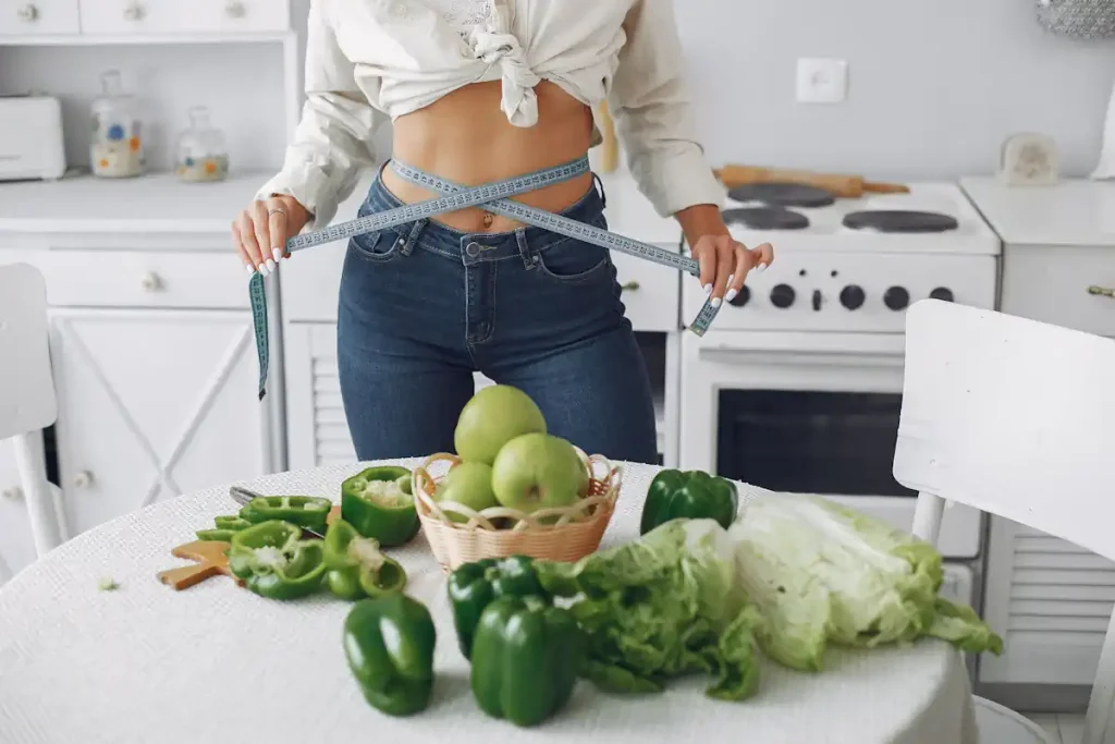 Une femme qui mesure son tour de hanche dans la cuisine devant une table avec plusieurs légumes verts