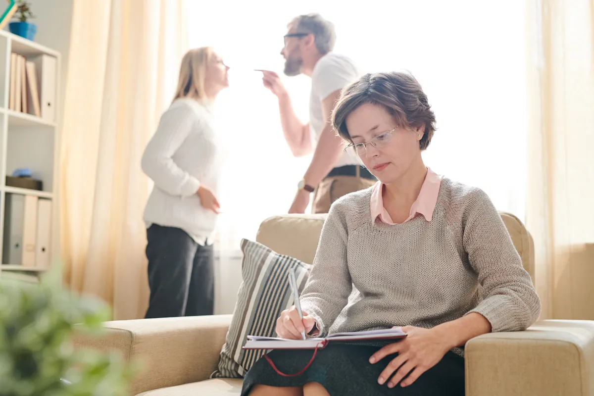 Une femme assise dans une chaise en prenant des notes avec derrière elle un homme et une femme qui se disputent