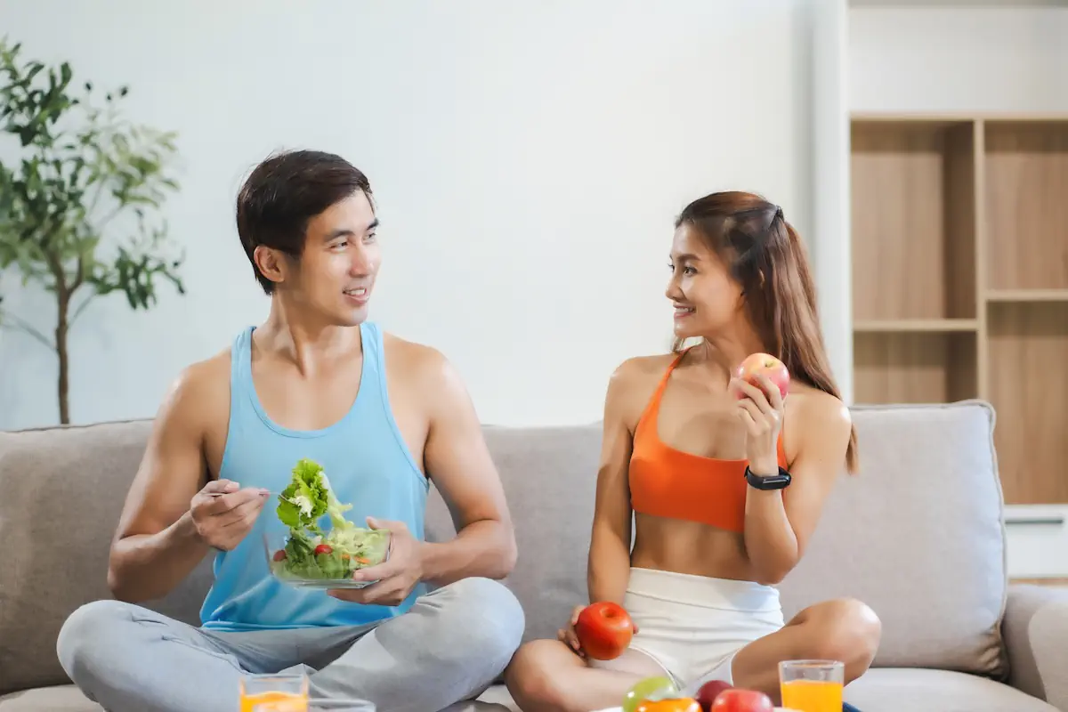Un couple en tenu de sport assis sur le canapé en tenant des légumes dans les mains