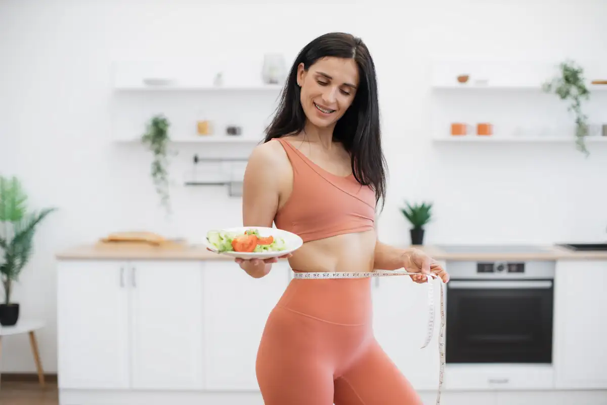 Une femme en teue de sport qui mesure sa talle avec un mètre ruban et qui tient une assiette de salade dans sa main