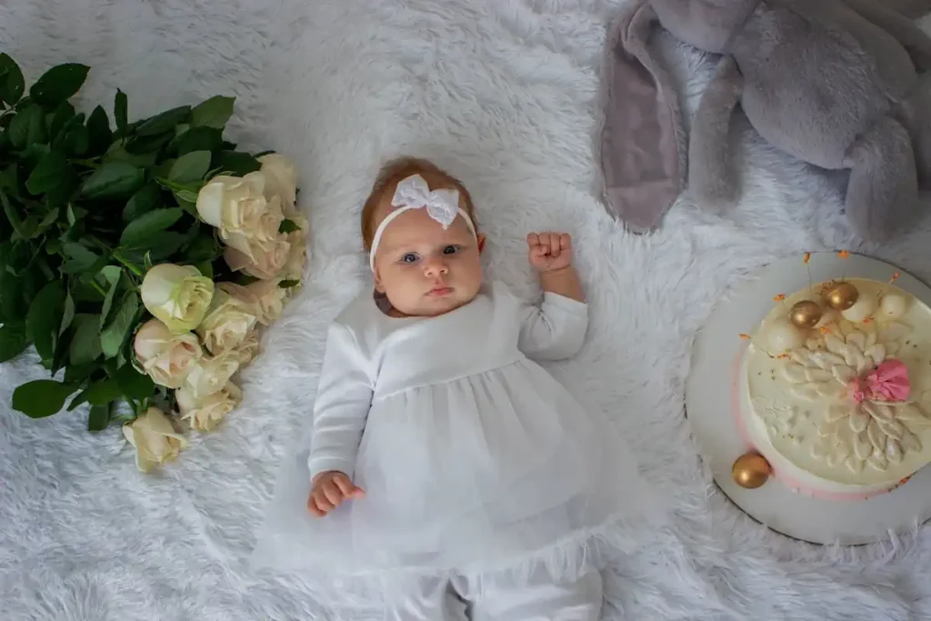 Une petite fille en robe blanche et avec un noeud blanc couché sur le dos à côté de fleurs et d'une peluche et d'un gâteau
