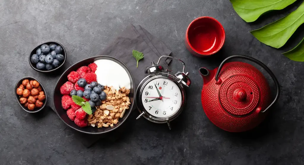 Une assiette de fruits et de graine posé à côté d'un réveil et d'une tasse et d'une cafétière rouge