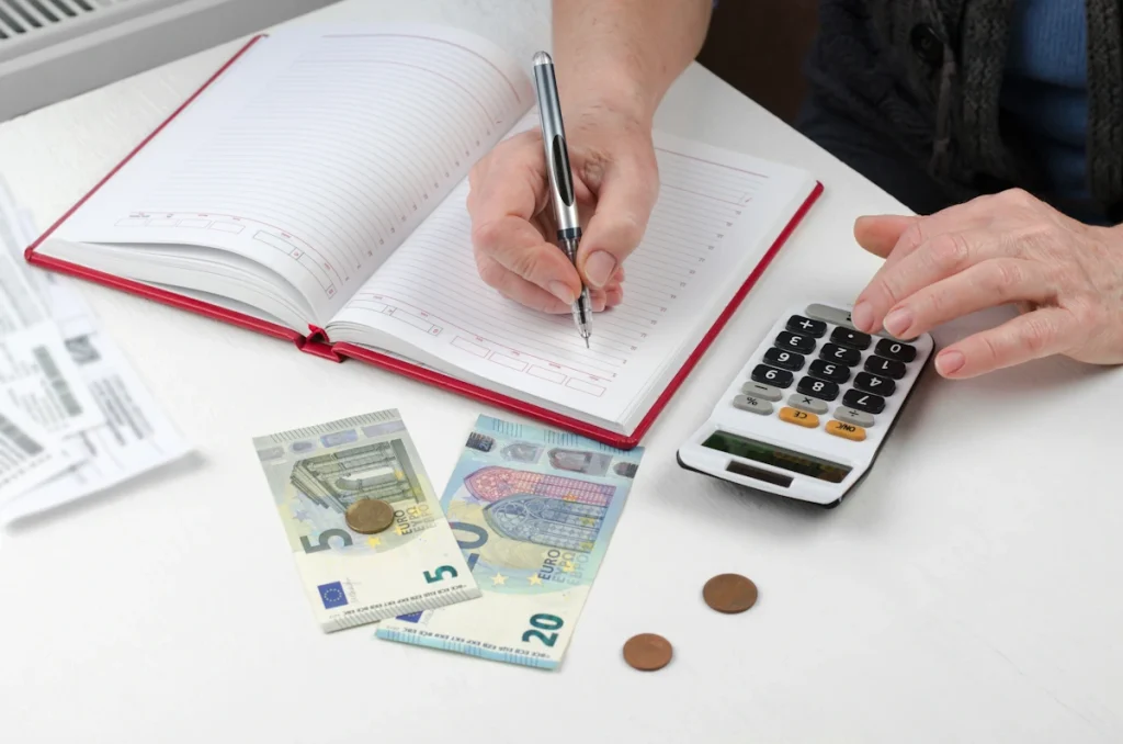 Une personne sur une machine à calculer et qui écrit sur un carnet avec de l'argent posé sur la table