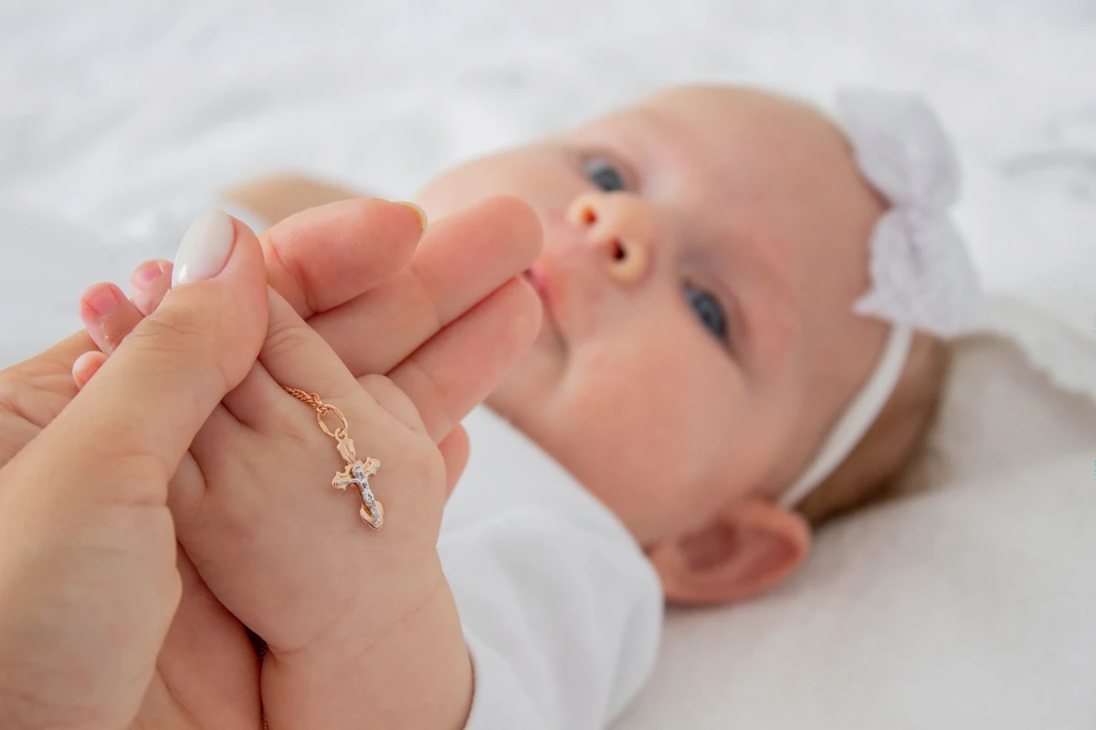 Un bébé avec un nœud blanc sur la tête avec une chaine avec un pendentif de croix dans sa main tenue par une femme