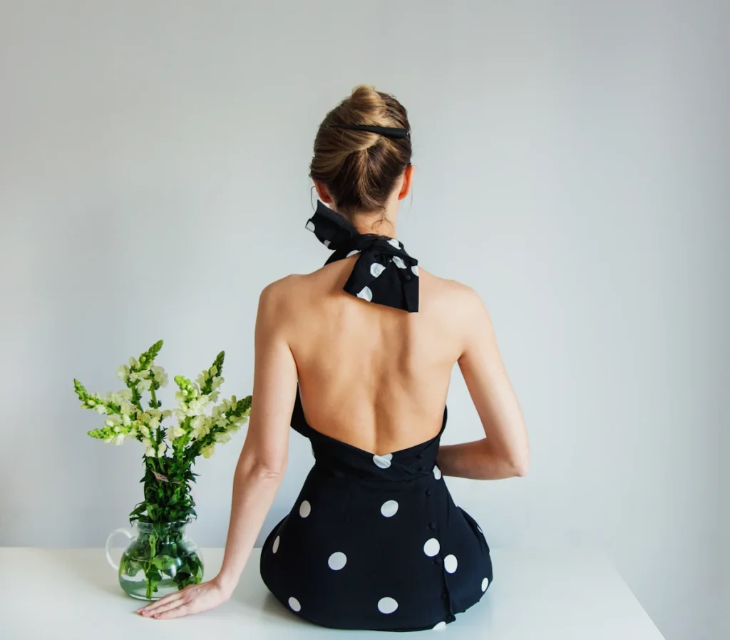 Une femme qui porte une robe noir à pois dos nu assise sur une table à côté d'une vase avec des fleurs blanches